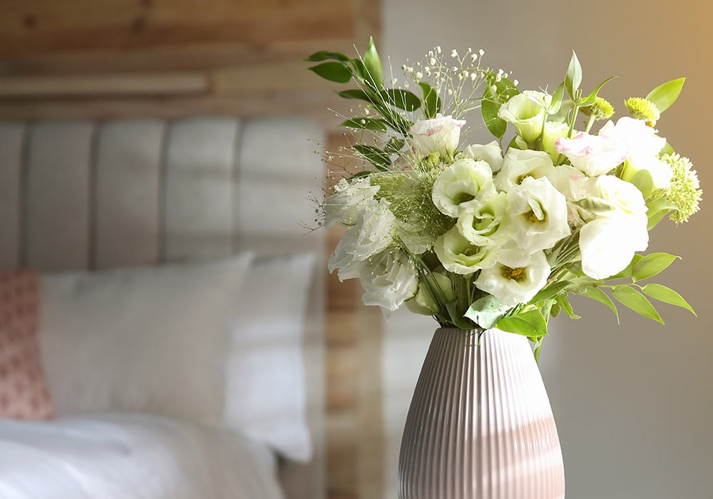 A vase of white and green flowers sits on a bedside table in a softly lit bedroom with a wooden headboard and white bedding.