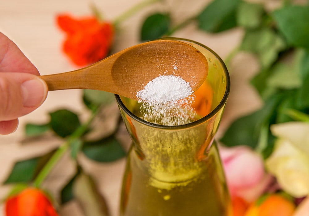 A wooden spoon with white powder is held over a glass bottle filled with liquid, surrounded by various colorful flowers in soft focus.