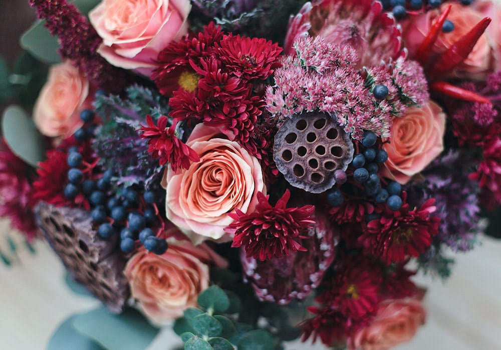 A bouquet of flowers displaying pink roses, crimson chrysanthemums, purple asters, berry clusters, and lotus seed pods, arranged together against a blurred green foliage background.