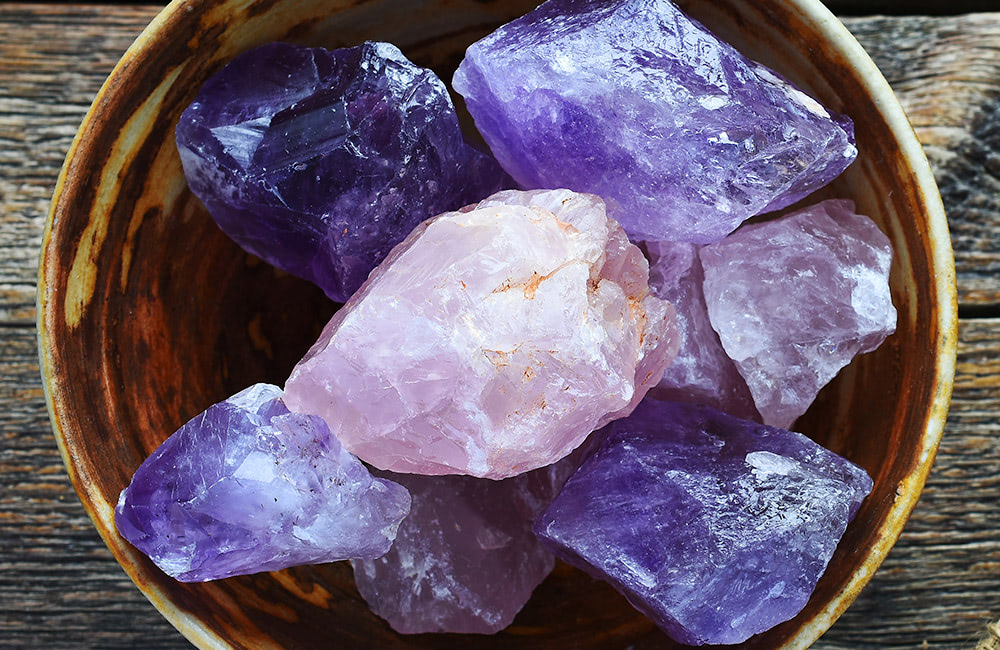 Raw amethyst crystals rest in a wooden bowl on a textured wooden surface.