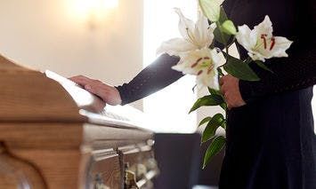 Person holding white lilies stands beside a wooden casket in a softly lit room, touching it with one hand.